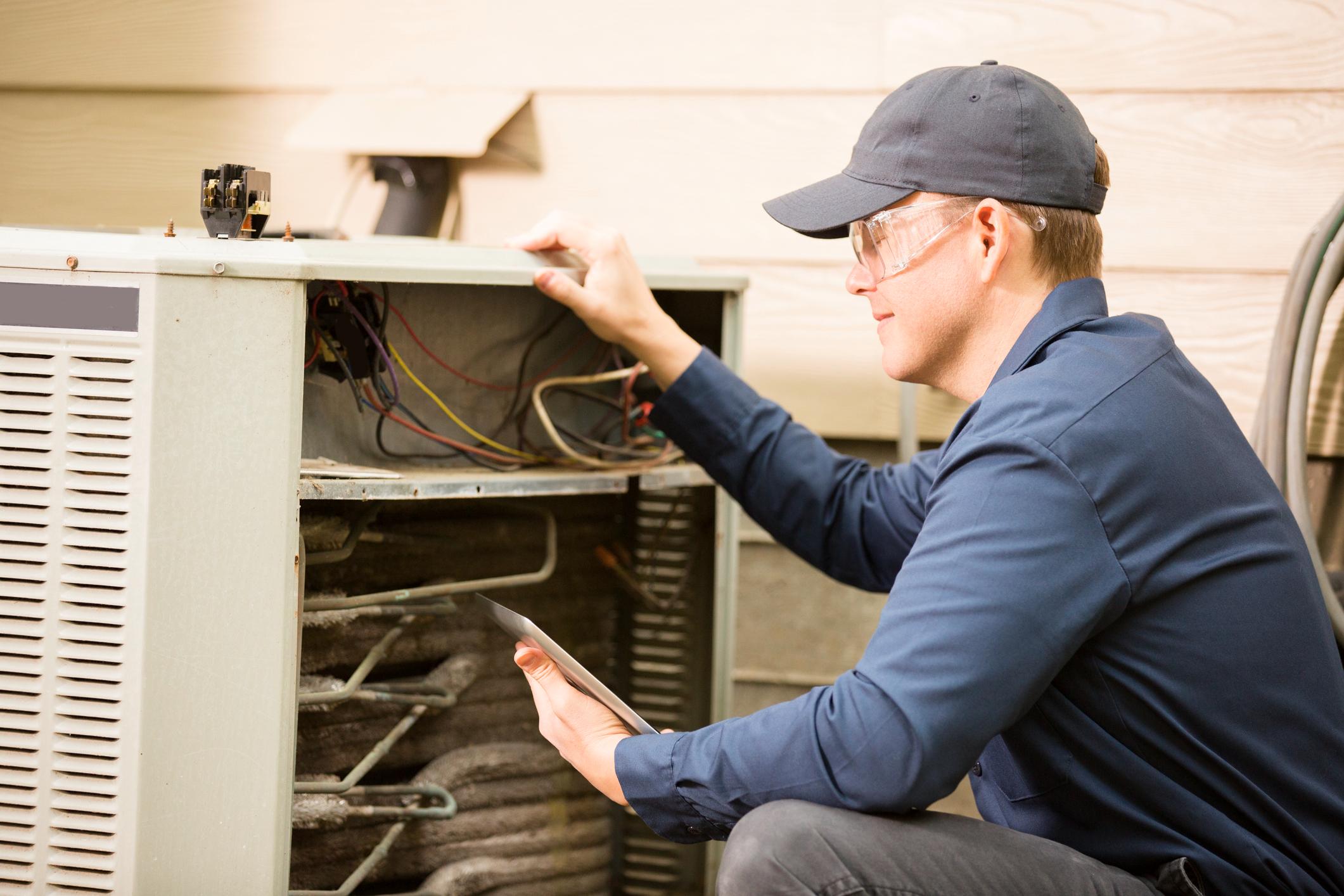 Repairmen works on a home's air conditioner unit outdoors. He is checking the compressor inside the unit using a digital tablet. He wears a navy blue uniform and his safety glasses.